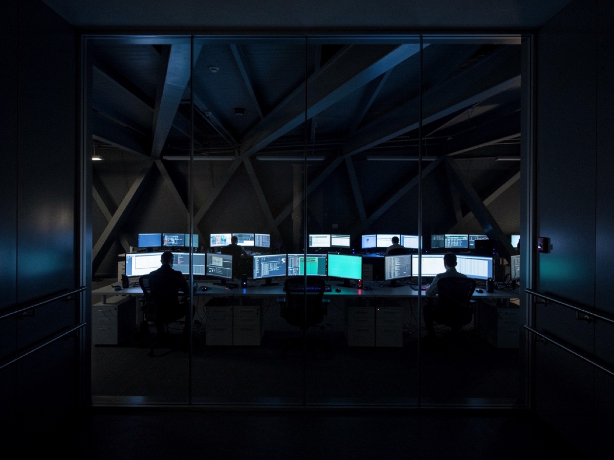 A security operations room seen at night through a glass partition, monitors lit inside